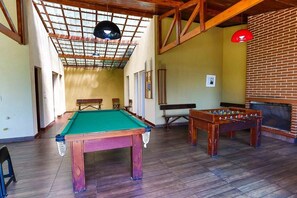 Une table de billard, une table de baby-foot et une cheminée dans une pièce dotée de poutres en bois et d’un plafond haut.