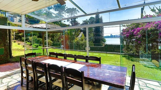 A conservatory with a table and chairs, large windows, and a view of a garden and water. - House facing the reservoir, in a gated community. (São Paulo)