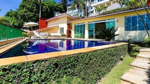 A swimming pool with lounge chairs, a red-roofed building, and a green fence. - House facing the reservoir, in a gated community. (São Paulo)