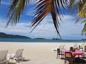 A beach with lounge chairs and a dining table, overlooking the sea and islands.