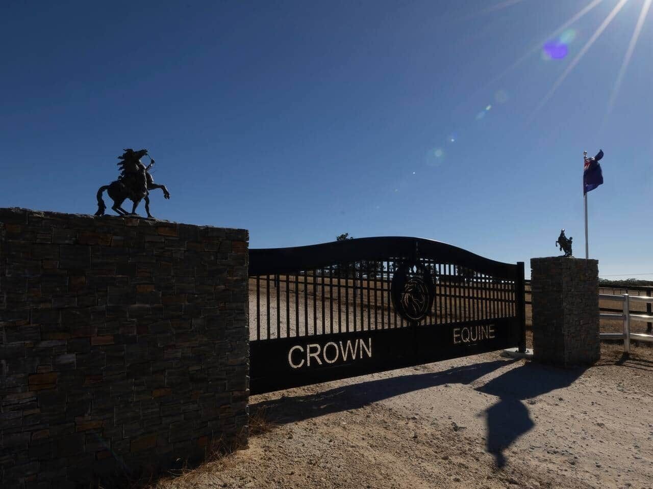 Ein verschlossenes Tor mit der Beschriftung 'CROWN EQUINE', eine Pferdestatue und eine Fahnenmasten.