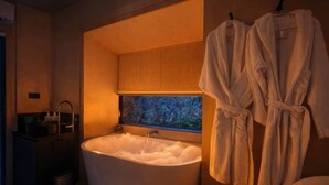 Un baño con una bañera independiente, una ventana que ofrece vistas a árboles y dos albornoces blancos colgados en un perchero.