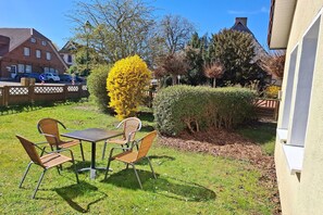 Une terrasse avec une table et des chaises, entourée de verdure et d’une haie.
