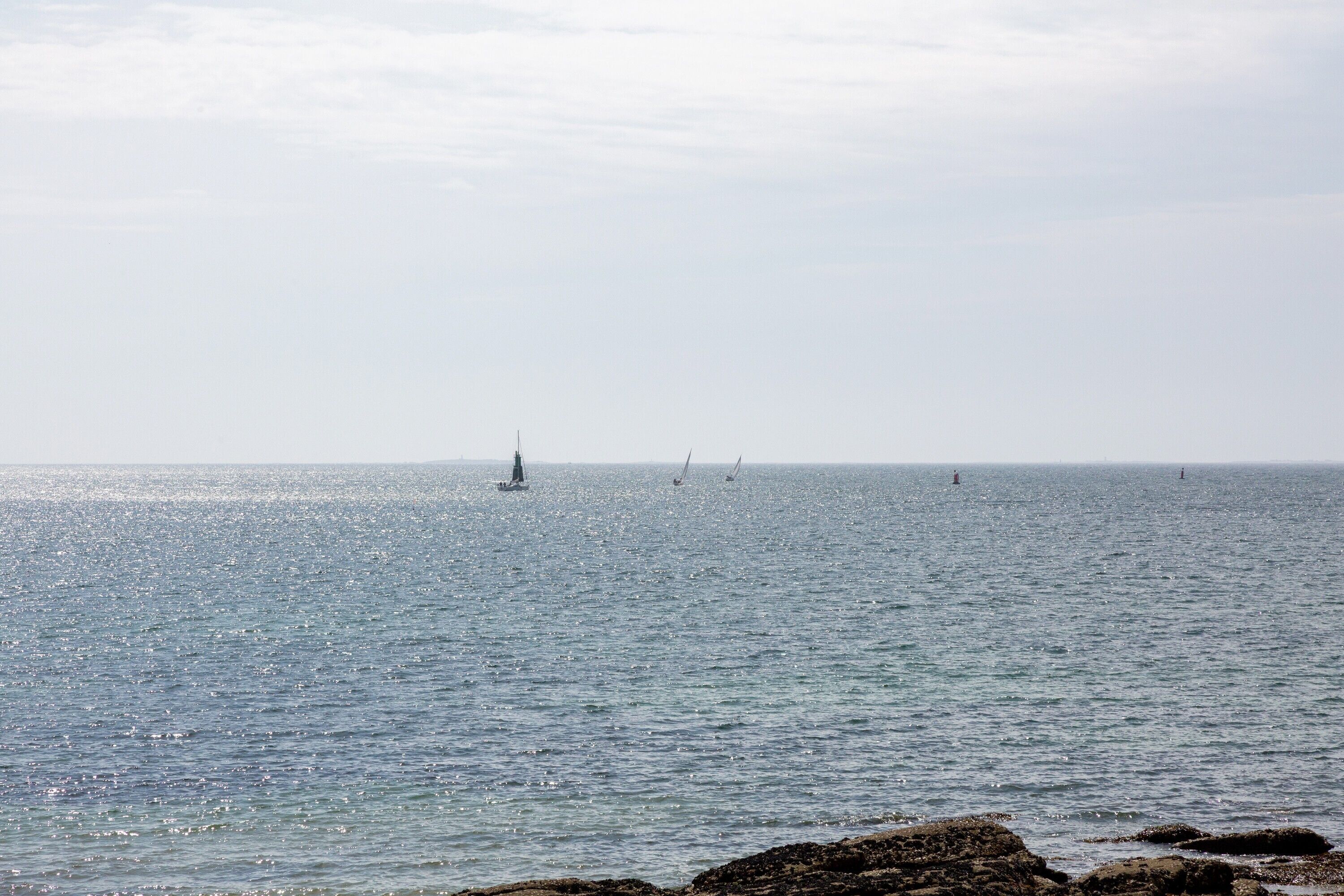 Mare calmo con alcune barche a vela, costa rocciosa e cielo sereno.