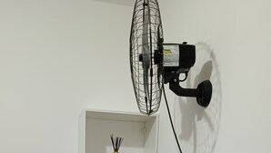 Une pièce avec un mur blanc, une étagère blanche, un ventilateur sur pied, une prise de courant et un lit de couleur sombre.