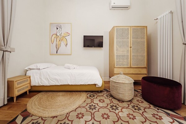 Ein Schlafzimmer mit Bett, Fernseher, Kleiderschrank, Sitzhocker aus Gewebe und Heizkörper.