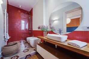 A modern bathroom with a red-tiled wall, a white sink, a large mirror, and a shower area. - Charming and modern 117 sq m apartment, accommodating up to 6 people, located on the first floor of a renovated building with an elevator. The property is strategically located, 700 meters from the historic Mercato del Capo and 500 meters from the Teatro (Palermo)
