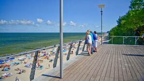 Une promenade en bois où l’on voit des gens se promener, une plage avec des parasols et des serviettes, et un ciel bleu dégagé.
