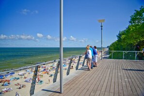 Ein hölzerner Steg mit Spaziergängern, ein Strand mit Sonnenschirmen und Handtüchern, und ein klarer, blauer Himmel.