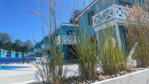 Un bâtiment à l’extérieur bleu et aux détails architecturaux blancs, entouré d’herbes hautes et surplombé d’un ciel bleu clair.