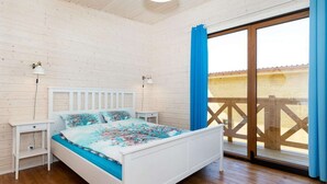 A bedroom with a white bed, two bedside tables, a nightstand, a window with blue curtains, and a wooden deck outside. - Comfortable holiday home (no. 6), summer pool (Rewal)