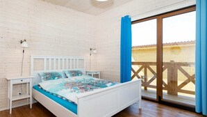 A bedroom with a white bed, a nightstand, a lamp, and a view of a wooden deck through a sliding glass door.