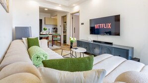 Moderni olohuone, jossa on sohva, vihreät koristetyynyt, pieni pyöreä pöytä ja seinälle asennettu televisio, jonka näytöllä näkyy teksti »NETFLIX».