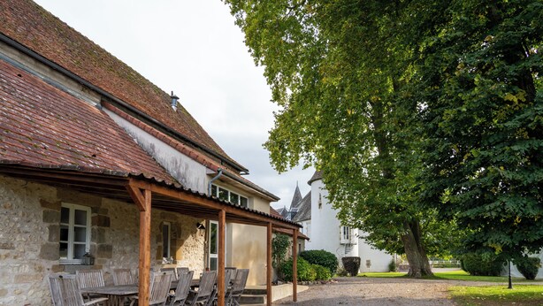 A stone building with a tiled roof, a wooden pergola with chairs, and a large tree in the foreground. - Le grand gîte du domaine d'Embourg (Souvigny)