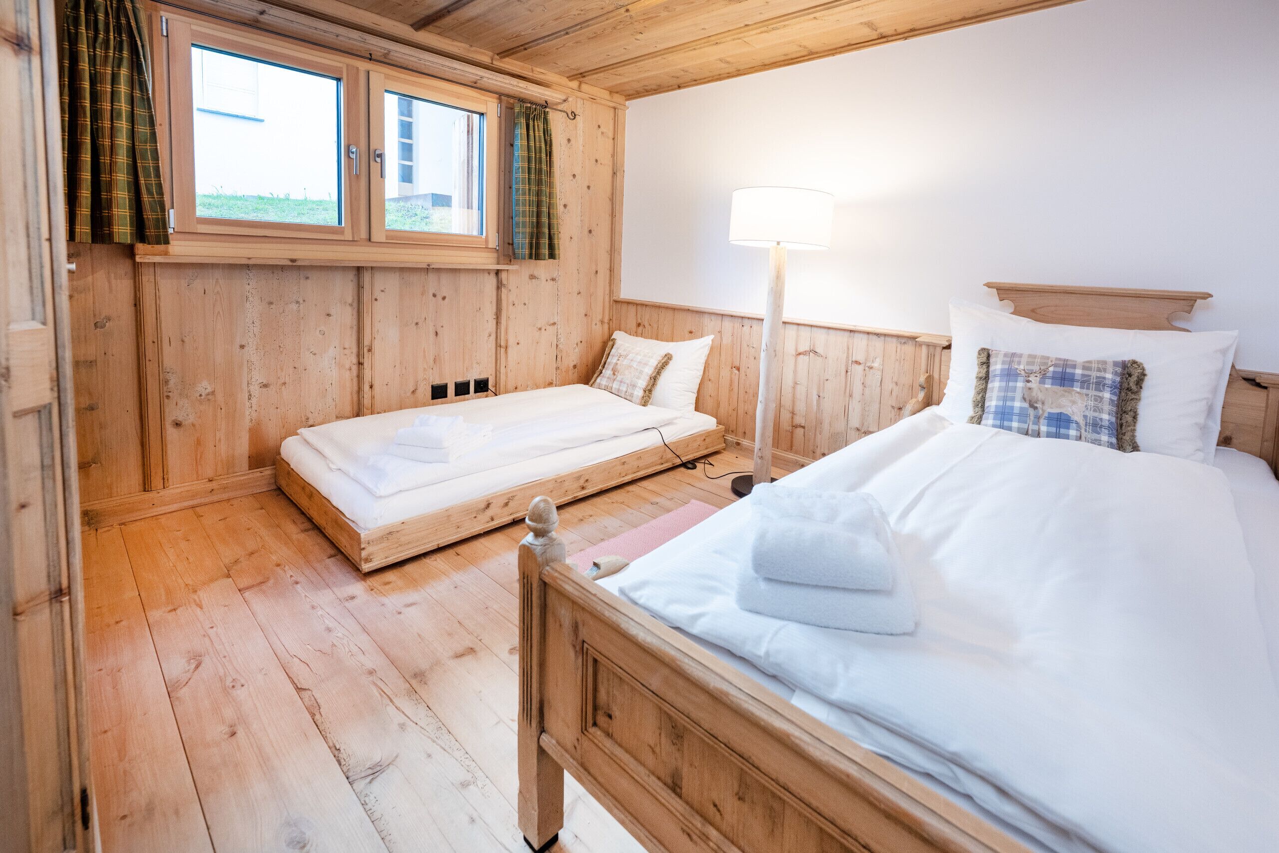 Ein Zimmer aus Holz mit einem Bett, einem zusätzlichen Bett, einem Fenster und einer Lampe.