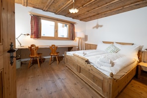 Un lit en bois avec du linge de lit blanc, un bureau avec une lampe, deux chaises en bois et une fenêtre avec des rideaux.