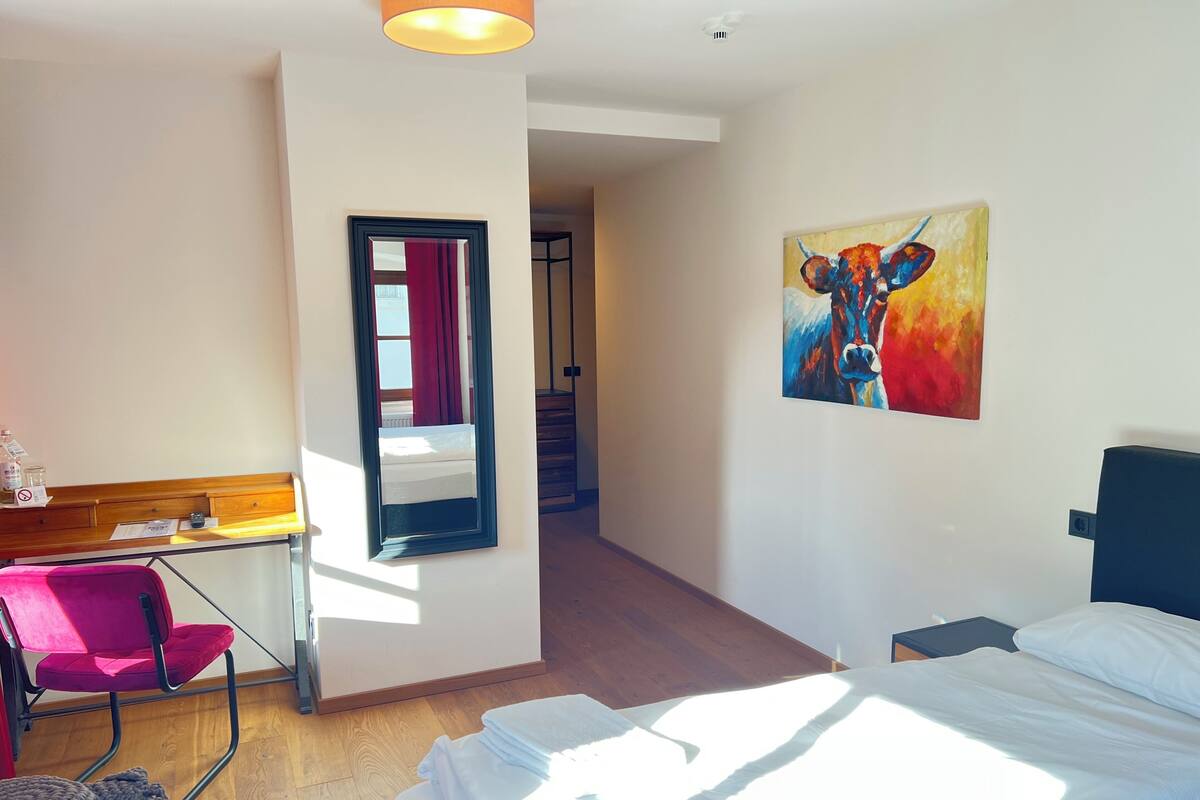 Ein Hotelzimmer mit einem Bett, einem Schreibtisch mit Stuhl, einem Spiegel, einem Bild an der Wand und einer Treppe im Hintergrund.
