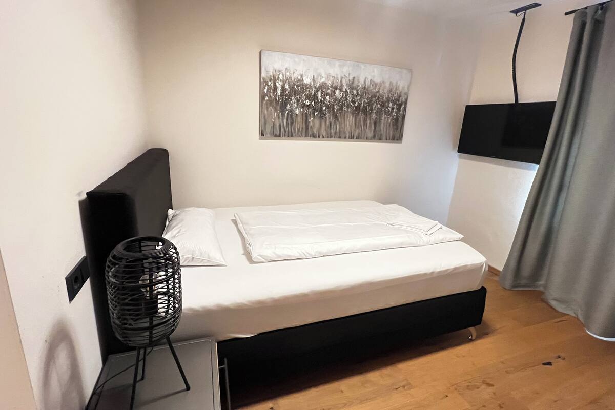 Ein Einzelbett mit schwarzem Kopfteil, ein an der Wand befestigtes Fernsehgerät, ein Gemälde und ein Standventilator.