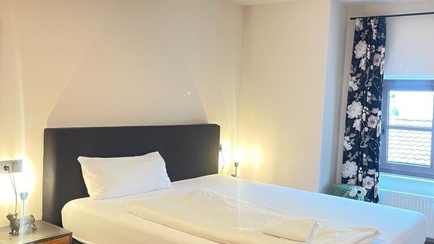 A neatly made bed in a room with a wooden floor, a nightstand with a lamp, and a window with floral curtains.