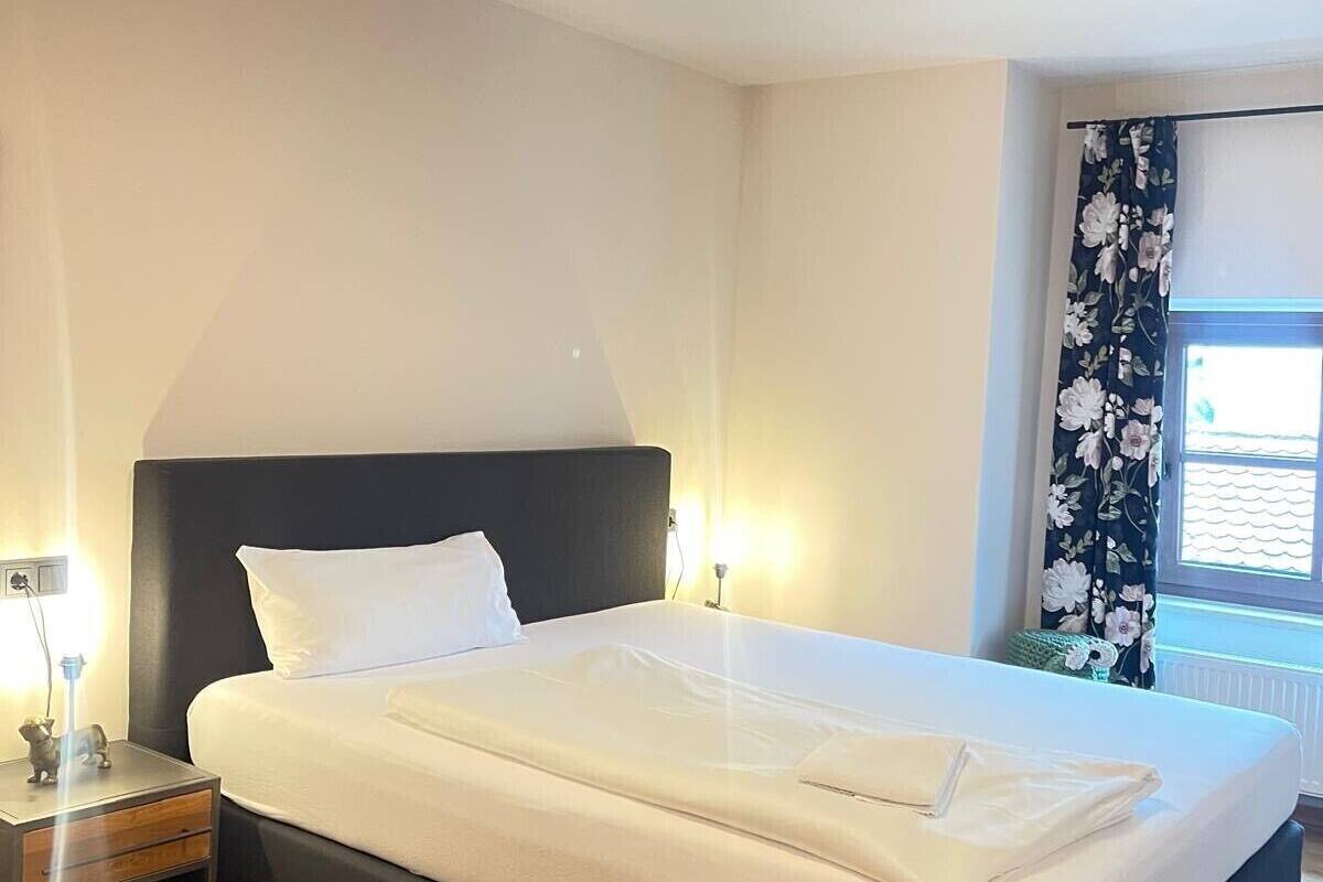 Ein ordentlich gemachtes Bett in einem Zimmer mit Holzboden, einem Nachttisch mit Lampe und einem Fenster mit floralem Vorhang.