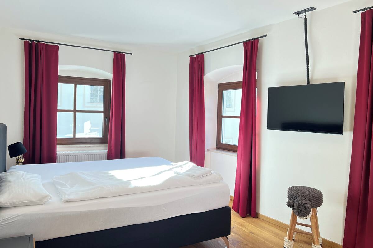 Ein Schlafzimmer mit einem Bett, einem Fernseher, einem Fenster und einem Hocker.