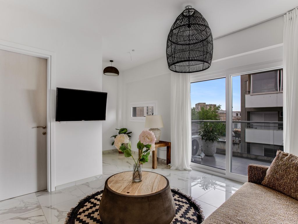 A modern living room with a flat-screen TV, a round wooden coffee table, a sofa, and a large window with a view of buildings.