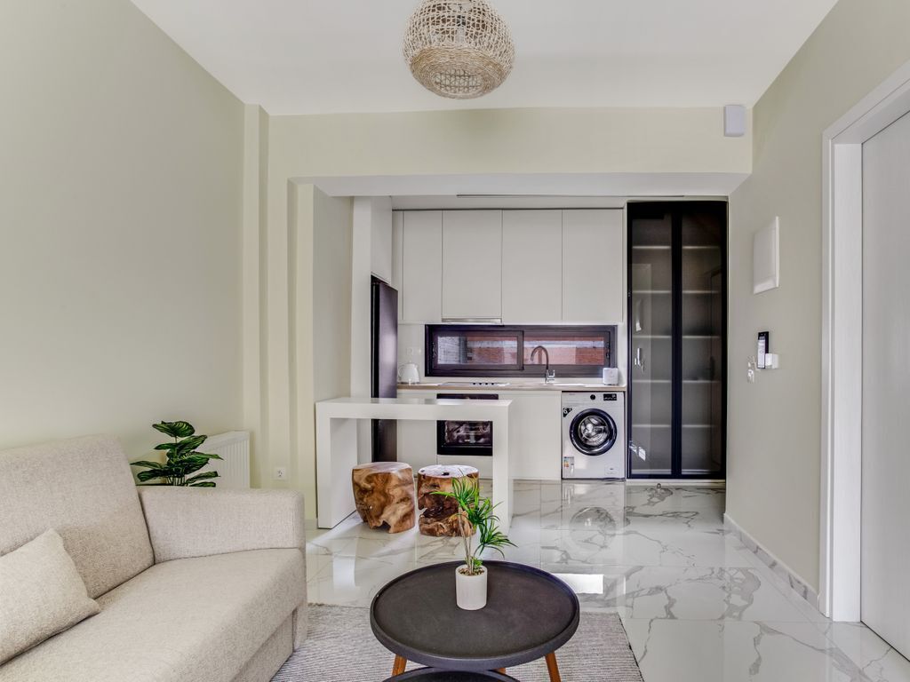 A modern living room with a sofa, coffee table, and decorative items. A kitchen with a washing machine and a TV is visible in the background.