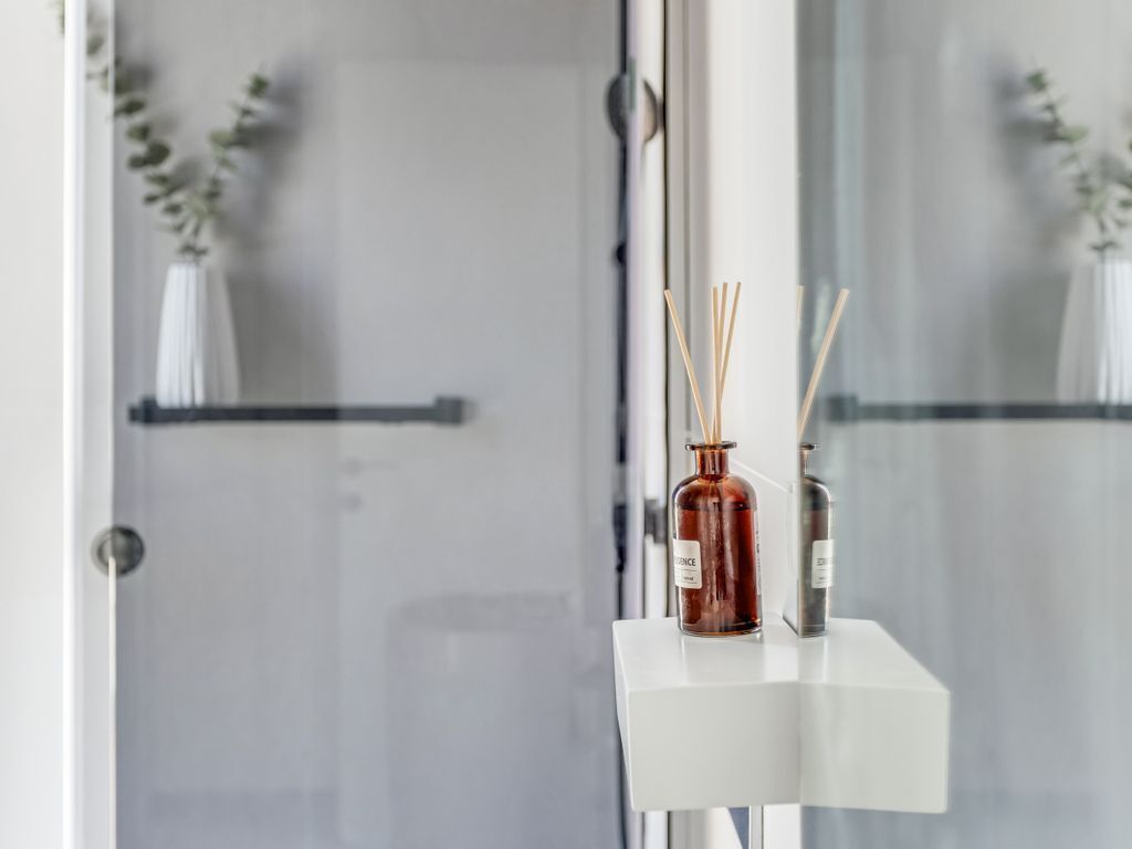 En duschvägg i glas med en vit hylla där två doftljus i form av rörformade ljushållare står.