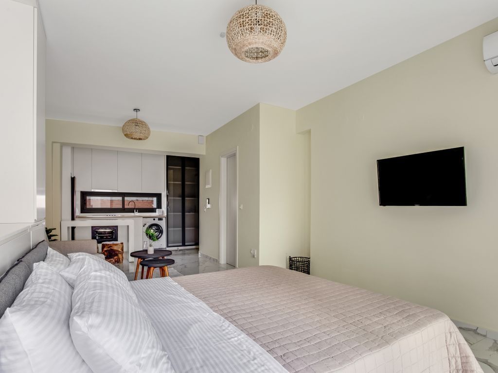 A bedroom with a bed, a television mounted on the wall, and a kitchenette in the background.