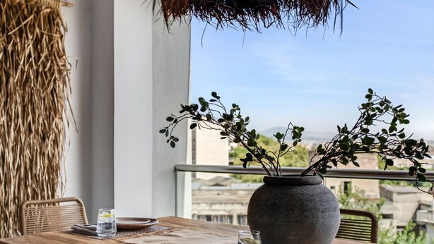 A dining table with a vase of greenery, plates, and glasses, set against a view of buildings. - Verde Premium Apartments (Thessaloniki)