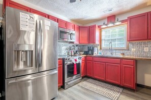 A kitchen with stainless steel appliances, red cabinets, a microwave, and a window with blinds. - Rustic 2BD Cabin w/ HUGE Yard | WiFi + Fire Pit (Gatlinburg)