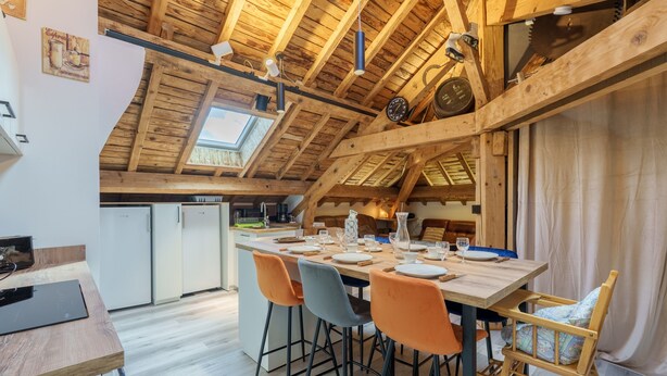 Comedor con mesa de madera dispuesta para cuatro personas, sillas alrededor, en una habitación con vigas de madera a la vista y tragaluz.
