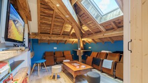 Habitación con techo de madera, tragaluz, televisor, mesa, sillas y una estantería.
