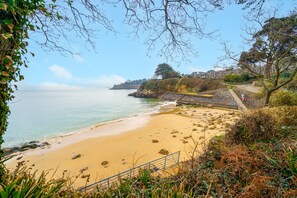 Uma praia costeira com areal arenoso, um afloramento rochoso e uma escadaria que desce até a praia.