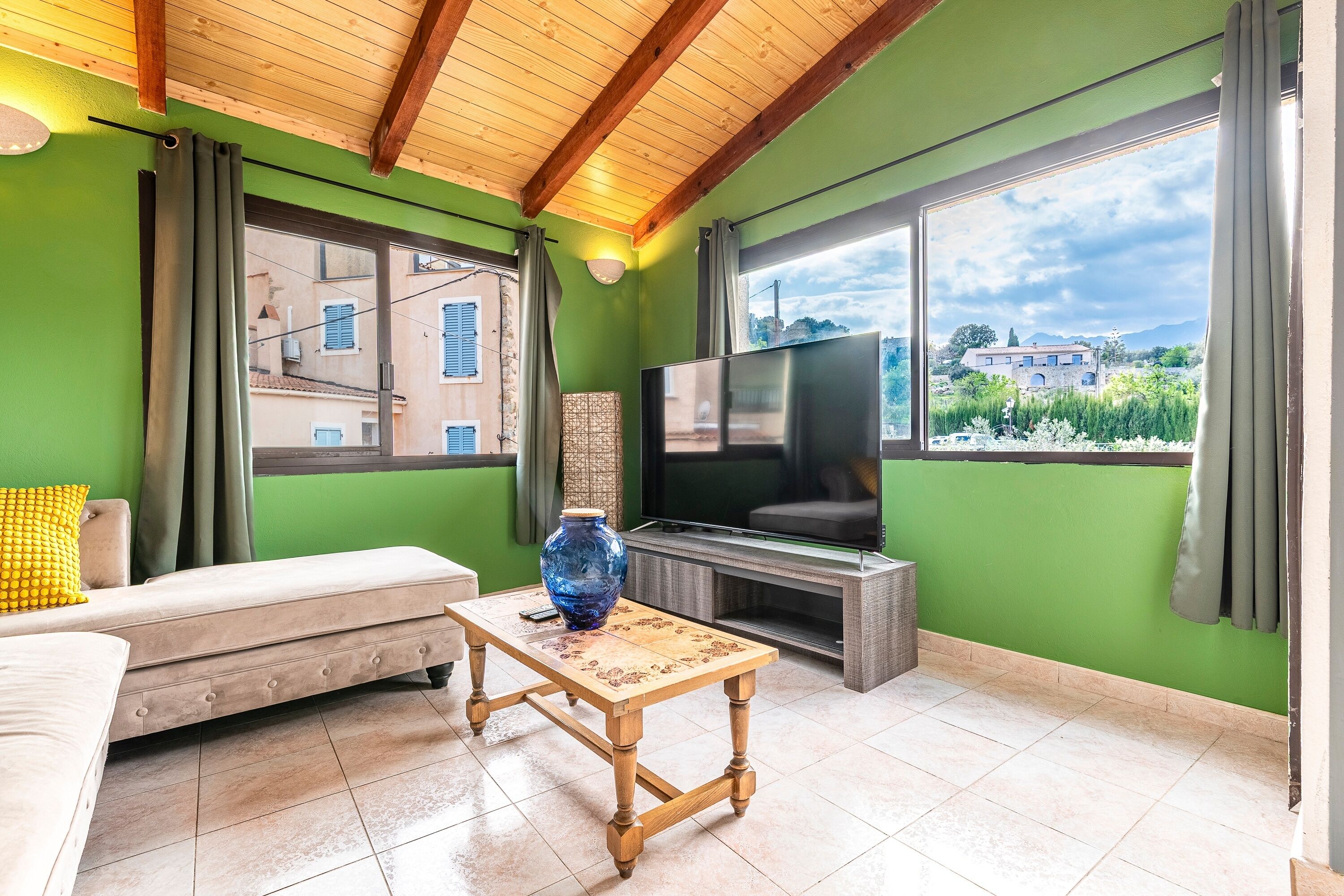 A living room with a sofa, a wooden coffee table, a TV, and a large window with a view of buildings and greenery.