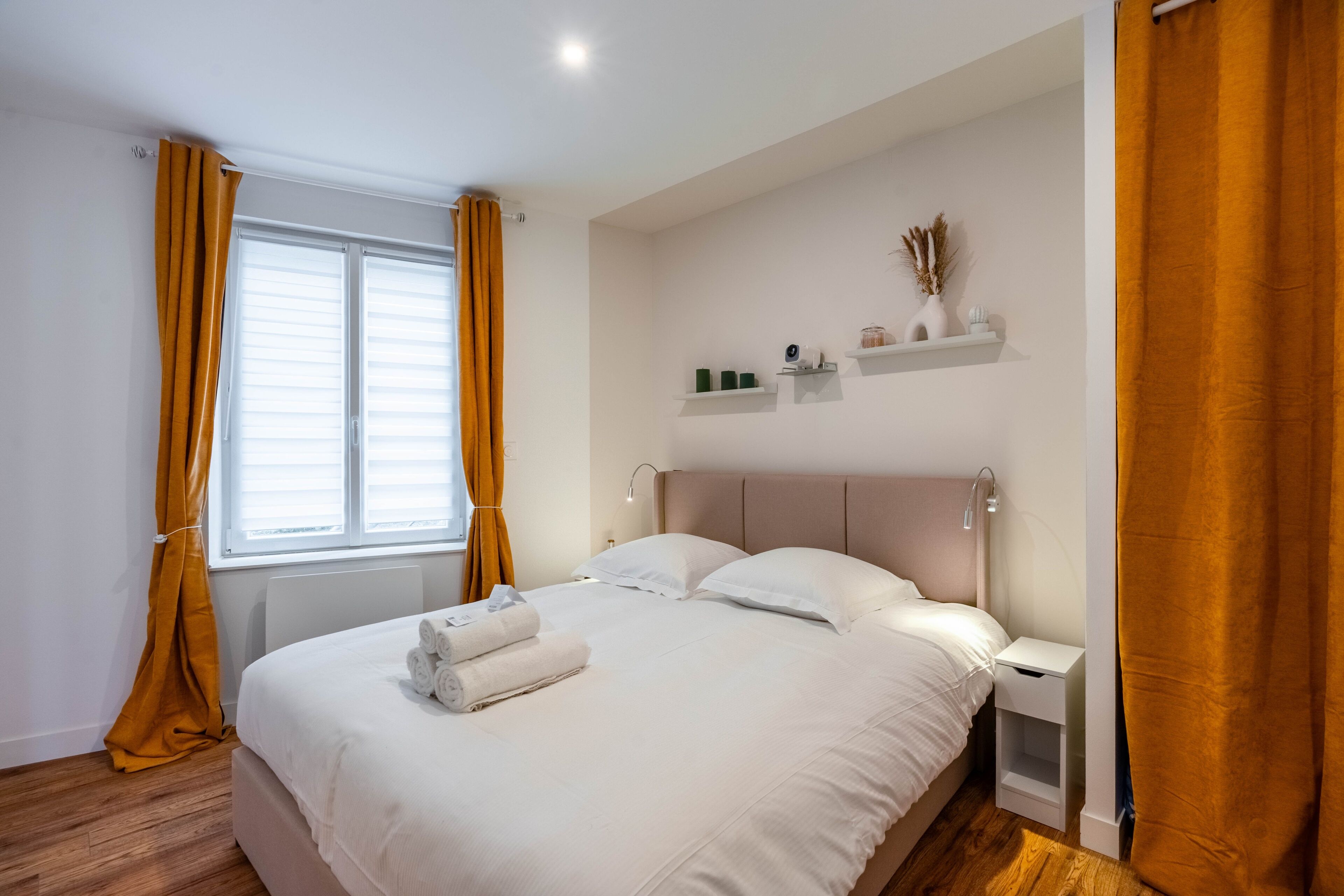 A neatly made bed with white linens, a bedside table with a lamp, and a window with curtains.