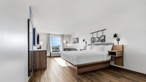 A modern hotel room with a wooden floor, a bed with white linens, a bedside table, a nightstand, a wall-mounted shelf, and a window with a view. - Sleep Inn & Suites Savannah Tennessee Riverfront (Savannah)