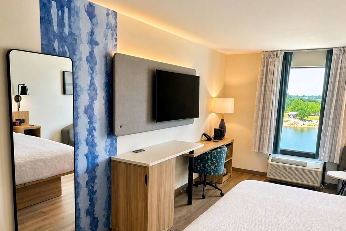 A hotel room with a bed, a desk, a TV, a mirror, a window with a view, and a lamp.