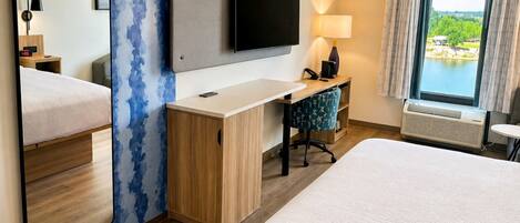 A hotel room with a bed, a desk, a TV, a mirror, a window with a view, and a lamp.