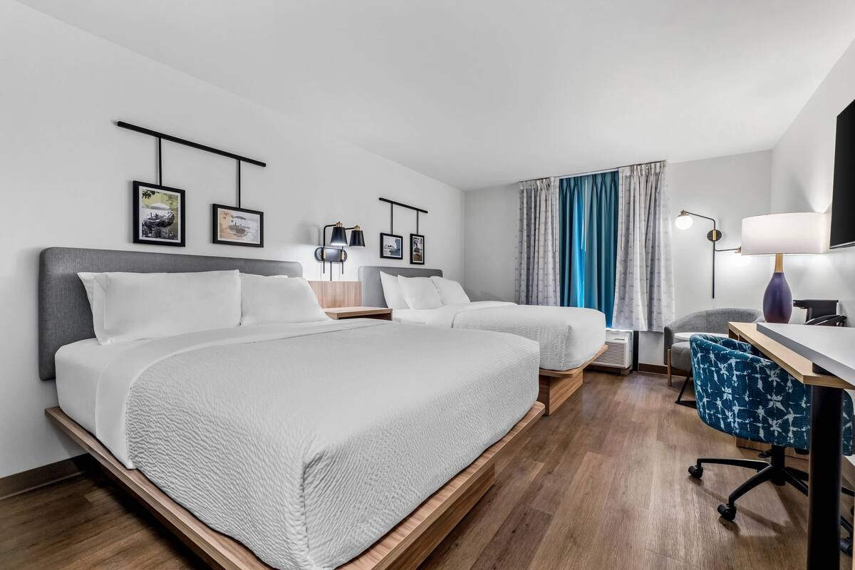 A hotel room with two beds, a desk with a chair, a TV, and framed pictures on the wall.