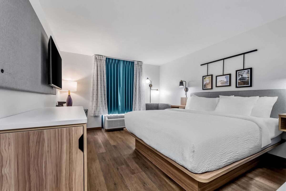 A modern hotel room with a large bed, a flat-screen TV, a desk with a lamp, and a wall-mounted picture.