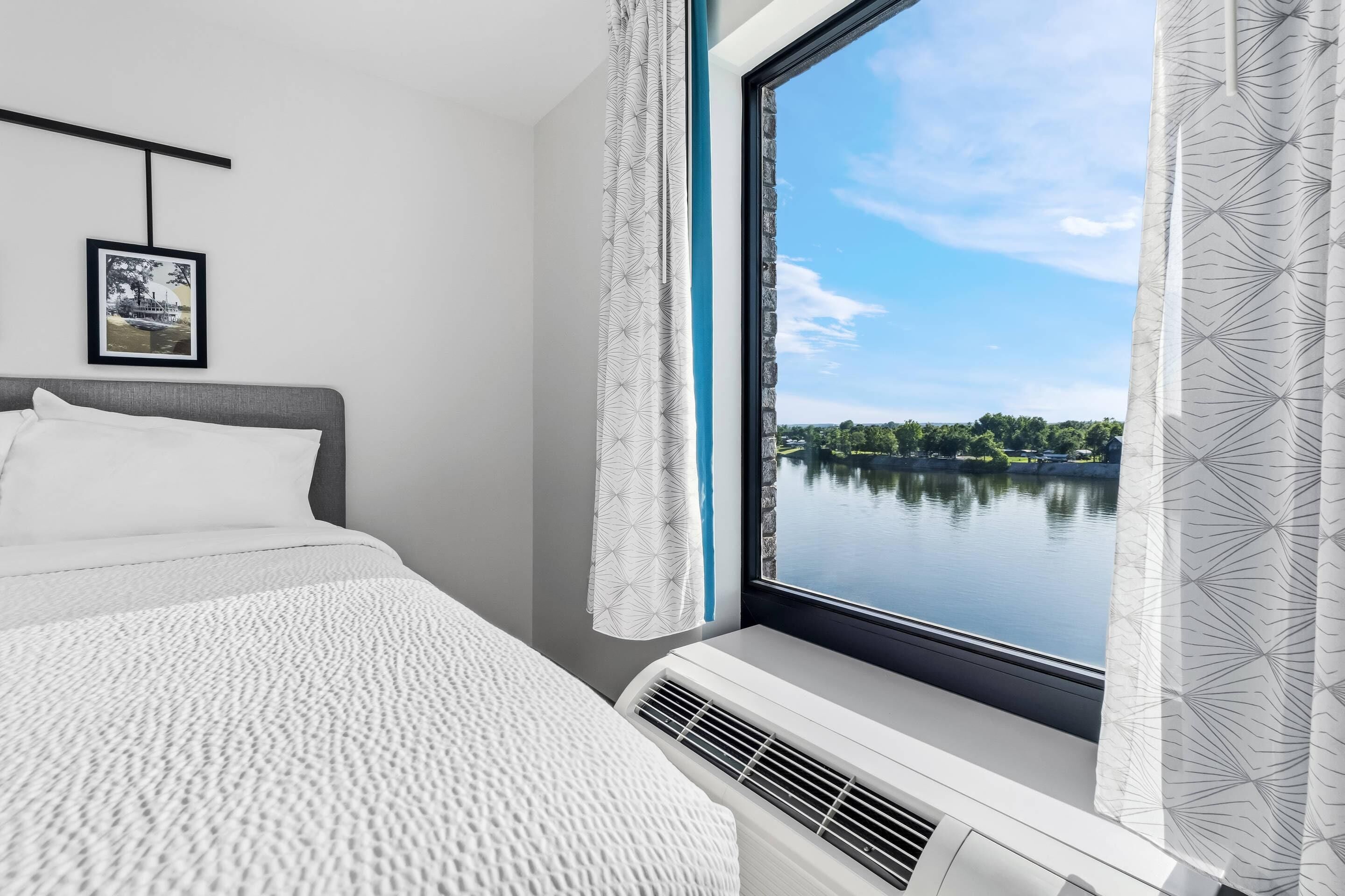 A bedroom with a bed, a window with curtains, a wall-mounted air conditioner, and a framed picture.