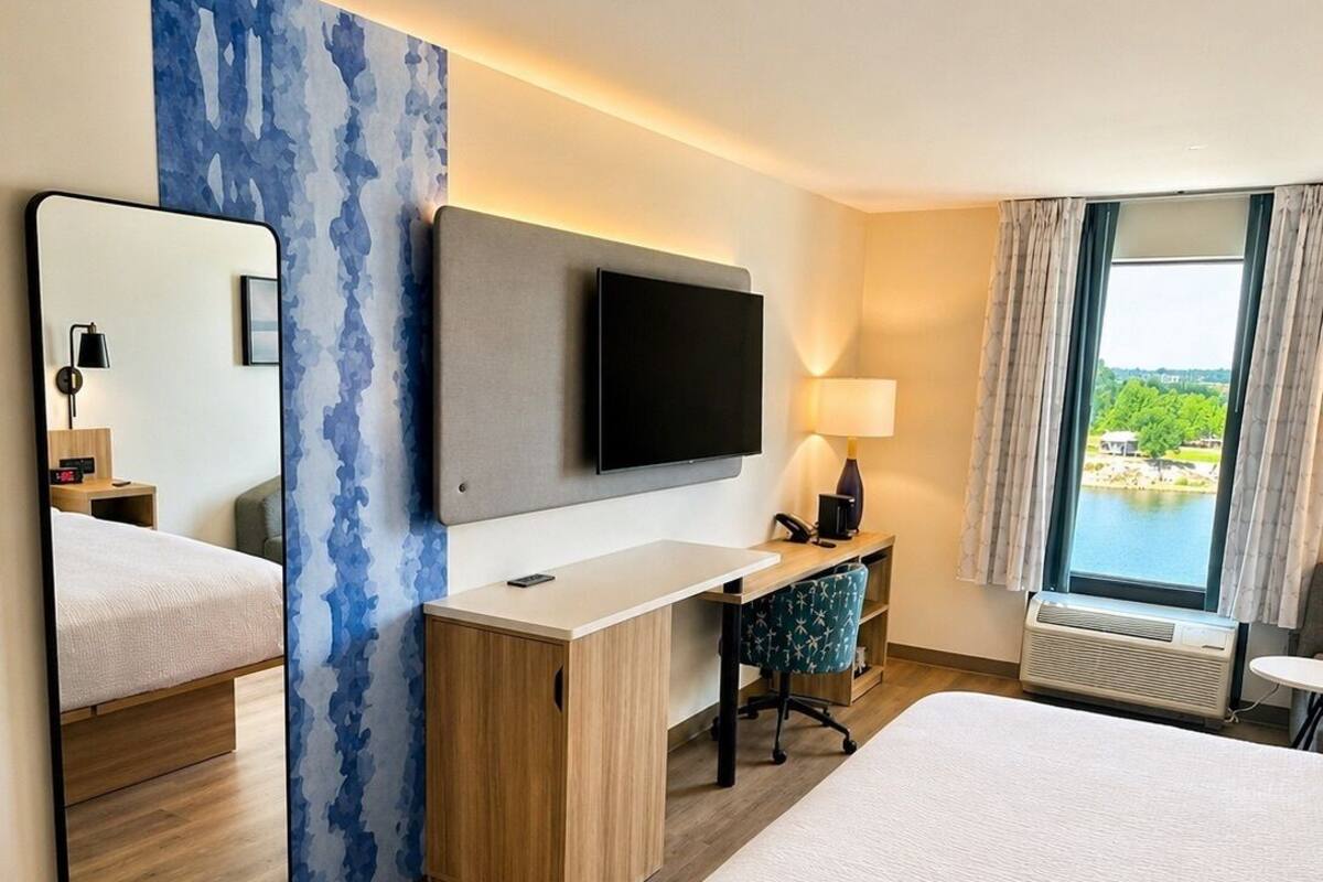 A hotel room with a bed, a desk with a flat-screen TV, a mirror, a lamp, and a window with a view.