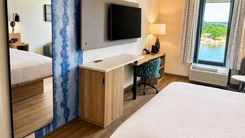 A hotel room with a bed, a desk with a flat-screen TV, a mirror, a lamp, and a window with a view.
