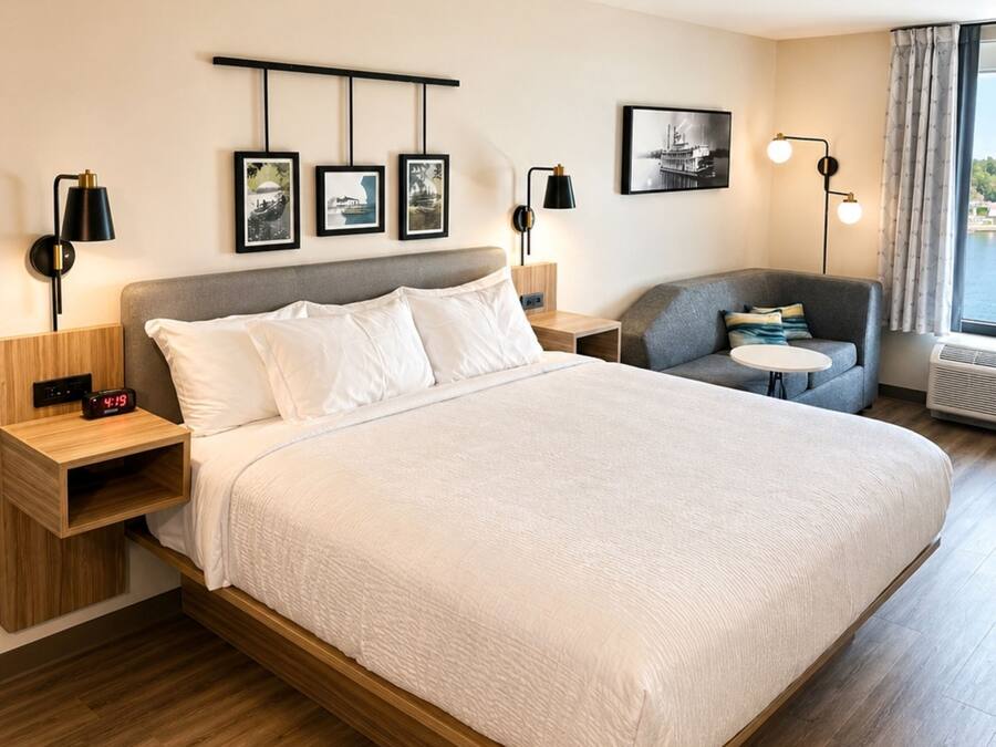Ein modernes Hotelzimmer mit einem großen Bett, Nachttischen, einer Sitzecke und Wandbildern, sowie einem Fenster mit Vorhängen.
