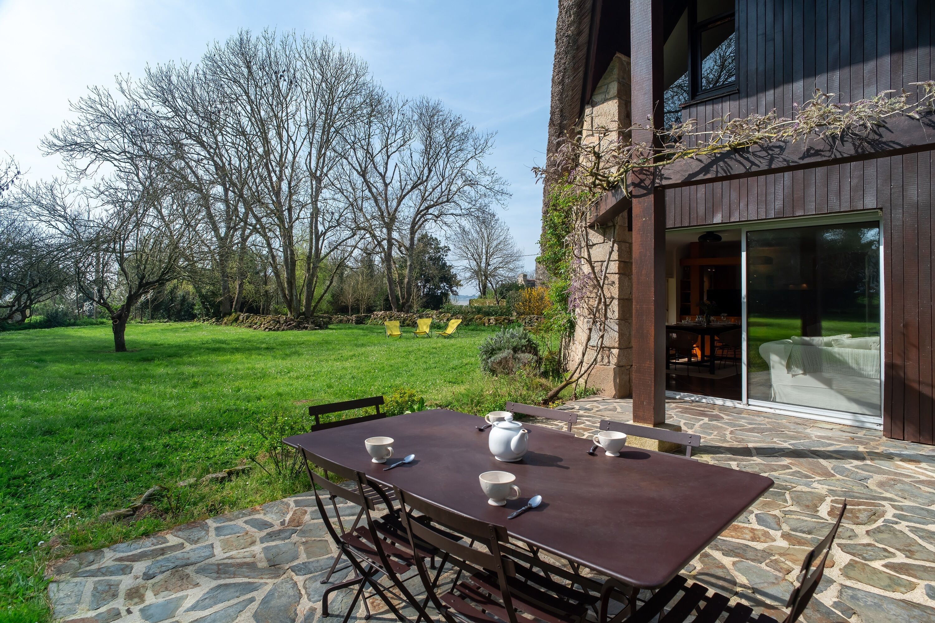 An outdoor patio with a stone table and chairs, a view of a grassy yard, and a building with large windows.