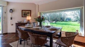 Zona pranzo con un tavolo grande, sedie in vimini, un orologio e vista su un giardino.