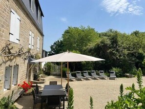 A courtyard with a table and chairs, a lounge area with sun loungers, and a building with stone walls and windows. - Plomarch Pella - à 300m de la plage (Douarnenez)