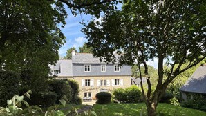 A two-story house with a stone facade, white shutters, and a visible front yard with a lawn and a small tree. - Plomarch Pella - à 300m de la plage (Douarnenez)