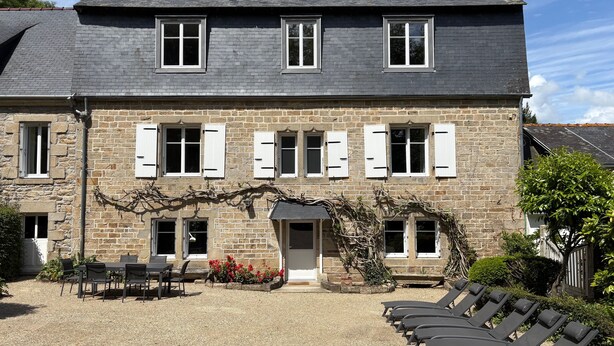 A stone building with a tiled roof, multiple windows with shutters, a gravel courtyard, outdoor seating, and a garden area. - Plomarch Pella - à 300m de la plage (Douarnenez)